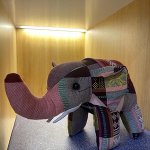 Op de afbeelding: Een stoffen olifantenbeeldje met een patchworkontwerp in bruin, roze, groen en paars. De olifant heeft een roze slurf, witte slagtanden en kleine zwarte ogen. Het beeldje is gemaakt van stof.
