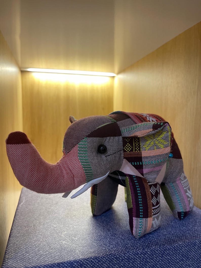 Op de afbeelding: Een stoffen olifantenbeeldje met een patchworkontwerp in bruin, roze, groen en paars. De olifant heeft een roze slurf, witte slagtanden en kleine zwarte ogen. Het beeldje is gemaakt van stof.