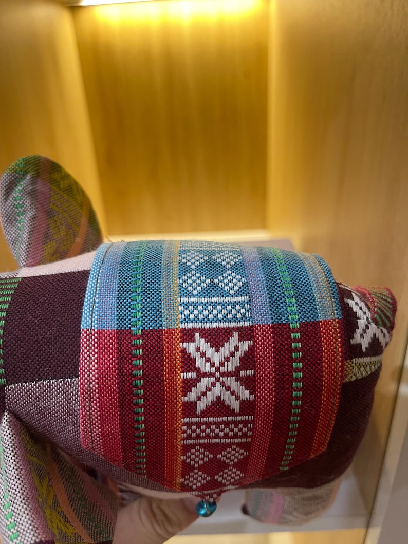 Op de afbeelding: Een kleurrijk, handgemaakt stoffen olifantenspeelgoed. De olifant heeft een patchwork-ontwerp met levendige rode, blauwe en witte patronen. Een kleine blauwe kraal hangt aan de onderkant. Het speelgoed is gemaakt van geweven stof.