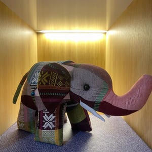 Op de afbeelding: Een patchwork olifantenbeeldje met een slurf, slagtanden en kleine oren. De olifant is gemaakt van verschillende stoffen met patronen in tinten bordeauxrood, groen, roze en cr&egrave;me. Het beeldje wordt op een blauw oppervlak weergegeven.