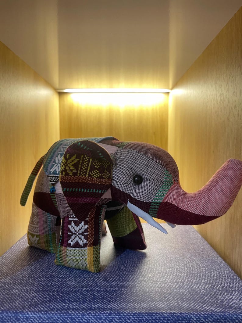 Op de afbeelding: Een patchwork olifantenbeeldje met een slurf, slagtanden en kleine oren. De olifant is gemaakt van verschillende stoffen met patronen in tinten bordeauxrood, groen, roze en cr&egrave;me. Het beeldje wordt op een blauw oppervlak weergegeven.