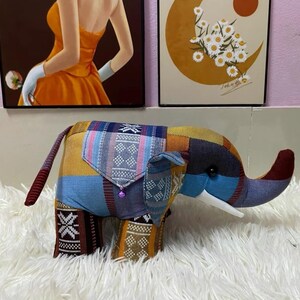 Op de afbeelding: Een kleurrijk patchwork olifantenbeeldje met een slurf, oren en staart. De olifant is gemaakt van verschillende stoffen met patronen in blauw, geel, oranje en bruin. Het heeft een klein zakje en een paarse kraal.