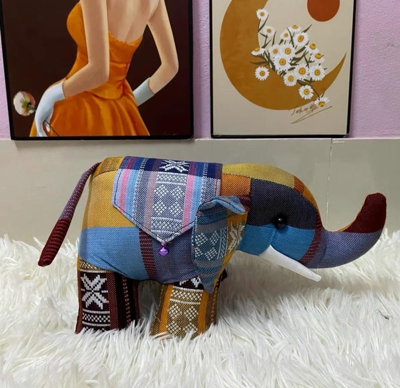 Op de afbeelding: Een kleurrijk patchwork olifantenbeeldje met een slurf, oren en staart. De olifant is gemaakt van verschillende stoffen met patronen in blauw, geel, oranje en bruin. Het heeft een klein zakje en een paarse kraal.
