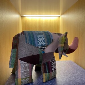 Op de afbeelding: Een patchwork olifantenbeeldje met een slurf en grote oren. De olifant is gemaakt van verschillende stoffen met patronen in groen, blauw, roze en bruin. Het wordt tentoongesteld op een blauw oppervlak.