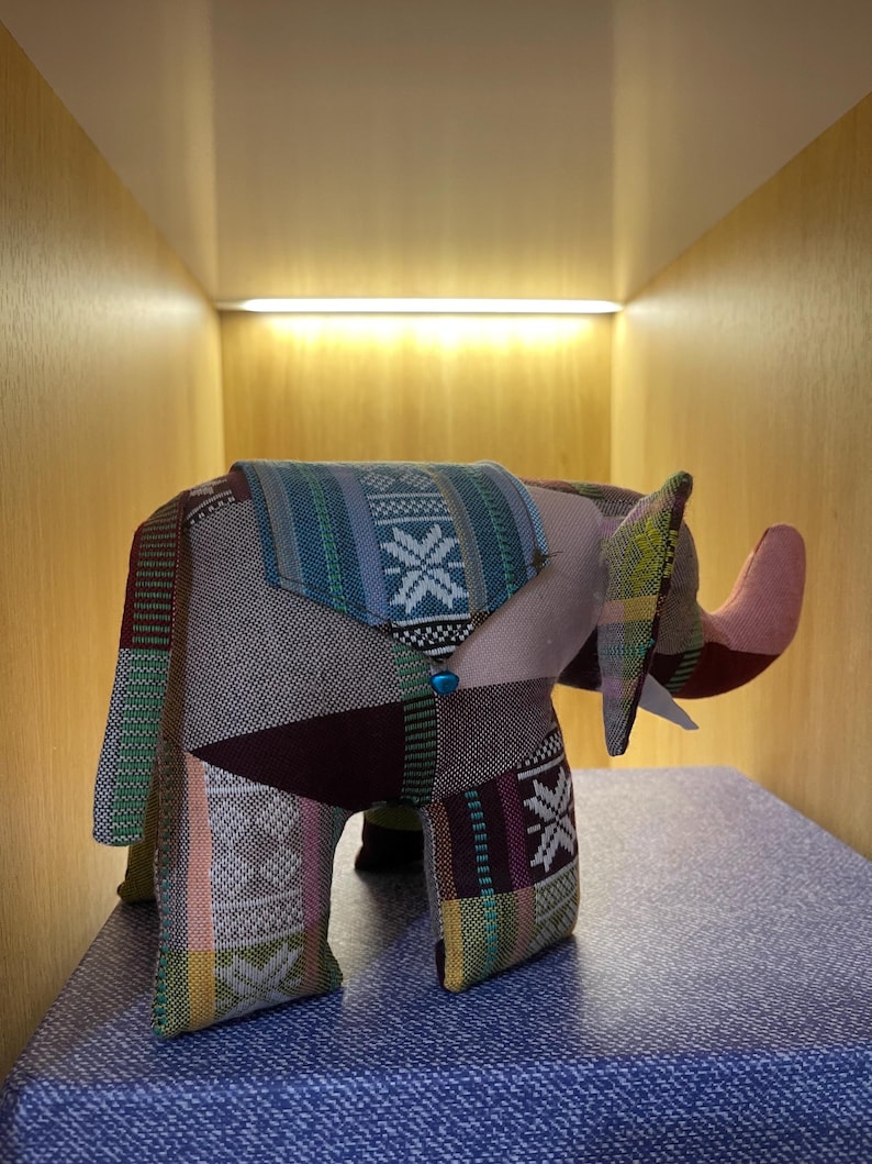 Op de afbeelding: Een patchwork olifantenbeeldje met een slurf en grote oren. De olifant is gemaakt van verschillende stoffen met patronen in groen, blauw, roze en bruin. Het wordt tentoongesteld op een blauw oppervlak.