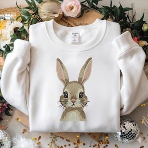May include: A white sweatshirt with a crew neck and long sleeves. The front features an embroidered bunny face with brown fur, pink inner ears, and black whiskers. The sweatshirt has a tag on the collar.