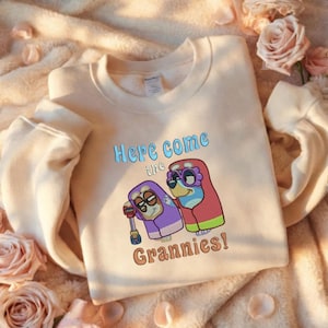 May include: A beige sweatshirt with the text "Here come the Grannies!" in blue and brown. The graphic features two cartoon characters in purple and red outfits with glasses. The sweatshirt is displayed on a soft, textured surface.