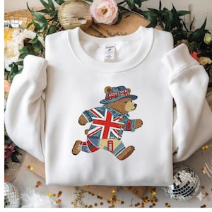 Könnte beinhalten: Weißes Sweatshirt mit einem gestickten Bären, der einen Union-Jack-Hut und eine Jacke trägt und rennt. Das Outfit des Bären enthält die Union-Jack-Flagge und eine rote Telefonzelle. Das Sweatshirt hat einen Rundhalsausschnitt und lange Ärmel.