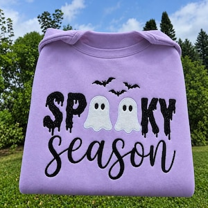 Pode incluir: Uma camisola de cor lilás claro com texto bordado em preto brilhante que diz SPOOKY season. O design inclui duas figuras de fantasmas brancos posicionadas entre as letras e três pequenos morcegos pretos bordados a voar acima.