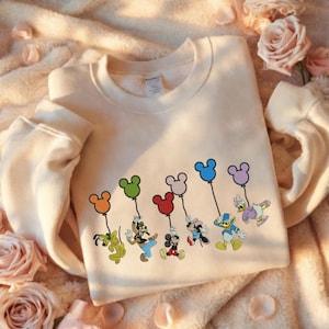 May include: Cream-coloured sweatshirt with embroidered Disney characters holding Mickey Mouse-shaped balloons in various colours. Characters include Pluto, Goofy, Mickey Mouse, Minnie Mouse, Donald Duck, and Daisy Duck.