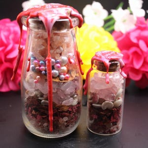 Puede incluir: Dos frascos de vidrio llenos de cristales, perlas y flores secas, sellados con cera roja. El frasco más grande mide aproximadamente 10 cm de alto y el más pequeño, unos 5 cm. Flores rosas y amarillas en el fondo.