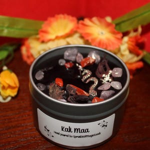 Puede incluir: Un recipiente de vela gris oscuro con un interior de cera negra, adornado con piedras decorativas, elementos botánicos secos y un dije de serpiente plateada. La etiqueta dice "Kali Maa".