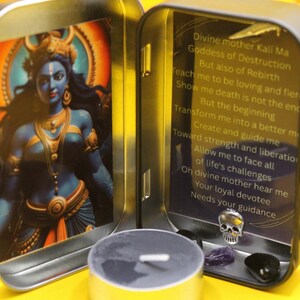 May include: A small, open metal tin featuring an image of a blue-skinned deity with gold accents. The tin also contains a small candle, a skull charm, and gemstones. Text on the inside panel reads "Divine mother Kali Ma Goddess of Destruction..."