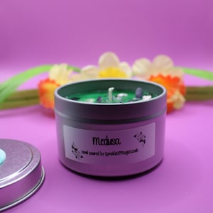 Puede incluir: Una lata de vela gris con un interior de cera verde, etiquetada como "Medusa". La vela está decorada con pequeñas piedras y una mecha blanca. La tapa está al lado de la vela. El fondo es de un morado vibrante.