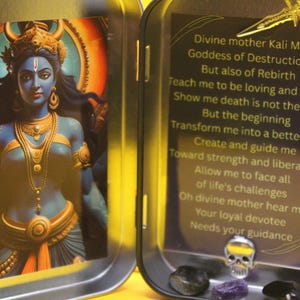 May include: A small, open metal tin with an image of a blue-skinned deity with gold jewelry and a crown. The right side of the tin has text about the goddess Kali Ma. The tin also contains small crystals and a skull.