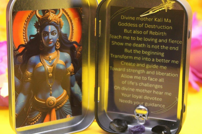 May include: A small, open metal tin with an image of a blue-skinned deity with gold jewelry and a crown. The right side of the tin has text about the goddess Kali Ma. The tin also contains small crystals and a skull.