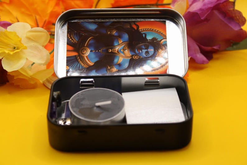 May include: A black metal tin with a hinged lid, open to reveal a religious image and small items. Inside are a tealight candle, a white square, and other small objects. The background is a bright yellow surface with flowers.
