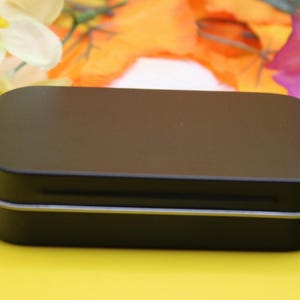 May include: A small, rectangular black tin with rounded corners. The tin has a silver trim around the edges. The background is a bright yellow with colorful flowers.