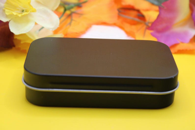 May include: A small, rectangular black tin with rounded corners. The tin has a silver trim around the edges. The background is a bright yellow with colorful flowers.