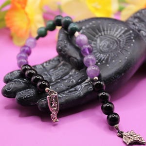 Könnte beinhalten: Ein Perlenarmband mit schwarzen, lila und grünen Edelsteinen. Das Armband ist mit silberfarbenen Charms verziert. Es wird auf einer schwarzen, handförmigen Skulptur vor einem rosa Hintergrund mit gelben und orangen Blumen präsentiert.