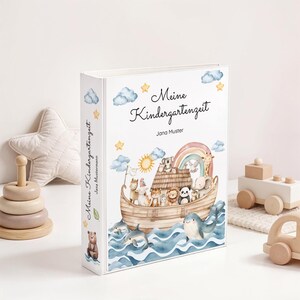 May include: A white binder with the German text "Meine Kindergartenzeit" and an illustration of an ark filled with animals, a rainbow, and a sun. The binder has a whimsical, watercolor style and is decorated with stars and clouds.