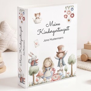 May include: A white binder with the words "Meine Kindergartenzeit" and "Jana Mustermann" printed on the cover. The cover features illustrations of characters from Alice in Wonderland, including Alice, the Mad Hatter, and the Cheshire Cat.