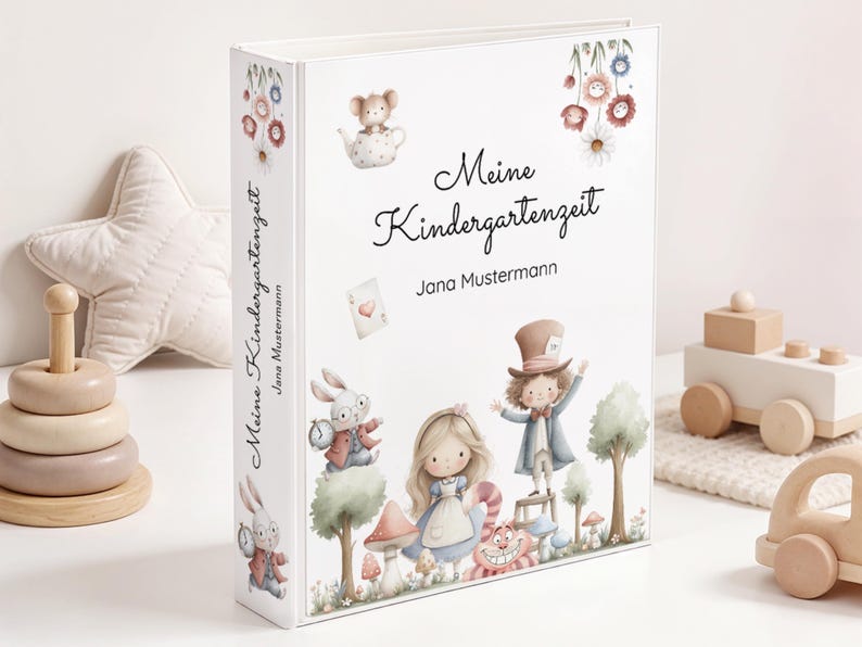 May include: A white binder with the words "Meine Kindergartenzeit" and "Jana Mustermann" printed on the cover. The cover features illustrations of characters from Alice in Wonderland, including Alice, the Mad Hatter, and the Cheshire Cat.