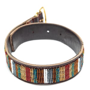 Puede incluir: Un collar para perros hecho a mano con una base de cuero marrón oscuro y una banda de cuentas. La banda de cuentas presenta rayas verticales en varios colores, incluyendo rojo, blanco, azul y dorado. El collar tiene una hebilla dorada.