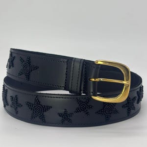 Puede incluir: Un cinturón de cuero negro con una hebilla dorada. El cinturón está decorado con cuentas negras en forma de estrella. Un accesorio elegante.