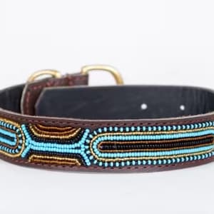 May include: Brown leather dog collar with a beaded design. The collar features a black interior, a gold-colored buckle, and is adorned with turquoise, gold, and black beads. The collar is designed for a pet.
