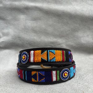 Puede incluir: Dos collares de perro de cuero negro con diseños de cuentas coloridas. El collar superior presenta cuentas naranjas, azules y blancas, mientras que el collar inferior tiene cuentas azules, rojas y amarillas. Ambos collares tienen una hebilla dorada.