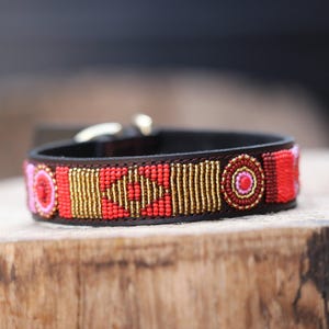 May include: A black leather dog collar with a beaded design. The collar features red, pink, and gold beads in a geometric pattern. The collar has a gold-colored buckle.