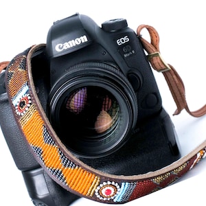 May include: A black Canon EOS 6D Mark II camera with a lens and a decorative beaded camera strap. The strap features a brown leather backing and is adorned with colorful beads in orange, red, blue, and white patterns.