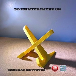 May include: A yellow, 3D-printed stand in an X-shape. The stand is made of a solid material and has a smooth finish. The text "3D PRINTED IN THE UK" is printed at the top of the image.