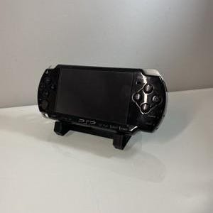 Op de afbeelding: Een zwarte PSP handheld gameconsole, rustend op een zwarte standaard. De console heeft een scherm, knoppen en een navigatiepad. De PSP is een draagbaar gaming-apparaat, ontworpen om onderweg games te spelen.