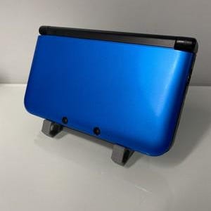 May include: A blue handheld games console with a black top and two small black buttons. The console is resting on a grey stand. The device has a glossy finish and a rectangular shape.