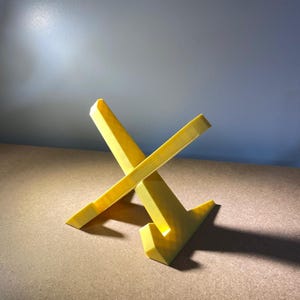 May include: A yellow, geometric stand with an angled design. The stand is made of a solid material and has a modern aesthetic. The stand is designed to hold a small object.