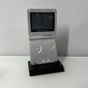 Può includere: Console portatile Game Boy Advance SP argentata, aperta e appoggiata su un supporto nero. La console presenta uno schermo e pulsanti di controllo. Il testo "GAME BOY ADVANCE SP" è visibile.
