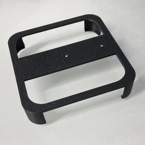 Può includere: Una staffa nera di forma quadrata con superficie strutturata. Presenta una sezione centrale rettangolare con due piccoli fori e angoli arrotondati. Il design è semplice e funzionale.