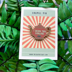 May include: A gold enamel pin shaped like a teapot with the text "I drink tea, darling" on a pink and white background. The pin is on a card that says "Enamel Pin" and "www.juliafrostart.com".