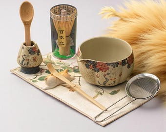Op de afbeelding: Een Japanse matcha theeceremonie set. De set bevat een keramische kom met bloemmotieven, een bamboe klopper, een bamboe lepel, een roestvrijstalen zeef, een keramische lepelhouder en een bamboe lepel. De set is geplaatst op een mat met bloemenpatroon.