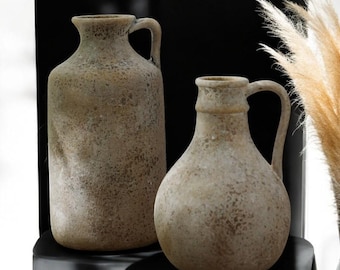 May include: Two beige ceramic vases with handles. The taller vase is cylindrical, while the shorter one has a rounded body. Both have a textured, speckled surface and are likely for home decor.