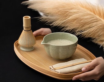 May include: A matcha tea set on a bamboo tray. The set includes a green ceramic bowl with matcha, a bamboo whisk, a small wooden spoon, and a linen cloth. The tray is oval-shaped and the background includes dried ornamental grass.