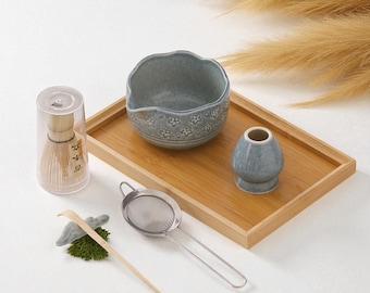 May include: A matcha tea set with a bamboo whisk in a clear case, a ceramic bowl with floral designs, a whisk holder, a stainless steel strainer, a bamboo scoop, and a wooden tray. The set is arranged on a white surface.