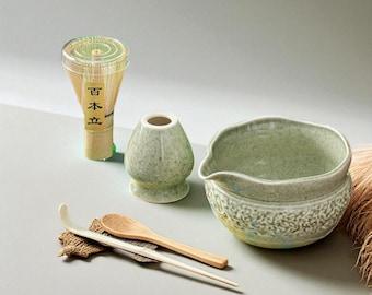 May include: A matcha tea set featuring a bamboo whisk, a ceramic bowl, a whisk holder, a scoop, and a spoon. The bowl and whisk holder are a speckled green. The whisk is in a clear container with Japanese text.