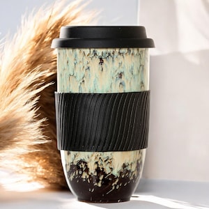 May include: A ceramic travel mug with a black lid and a black textured band. The mug has a unique glaze with shades of cream, teal, and brown. The mug is designed for hot or cold beverages.