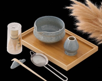 May include: A matcha tea set with a ceramic bowl, whisk, whisk holder, and a bamboo tray. The set includes a stainless steel strainer and a bamboo scoop. The bowl and whisk holder are a speckled blue-gray color. The whisk is in a clear plastic container.