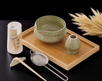 May include: A matcha tea set on a wooden tray. The set includes a green ceramic bowl, whisk holder, and a bamboo whisk in a clear container. Also included is a metal sieve and a bamboo scoop. The set is arranged on a wooden tray.