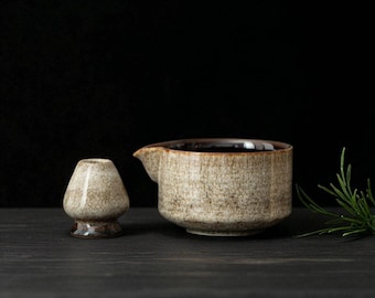 Puede incluir: Un juego de té matcha de cerámica con un batidor y un cuenco vertedor. El cuenco tiene un pico y un acabado texturizado y moteado en tonos marrones y beige. El batidor es pequeño y redondeado.