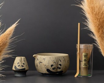 May include: A ceramic matcha tea set featuring a panda design. The set includes a bowl, a whisk, a whisk holder, and a small cup. The bowl and cup have panda illustrations. The whisk is in a clear container with Japanese text.